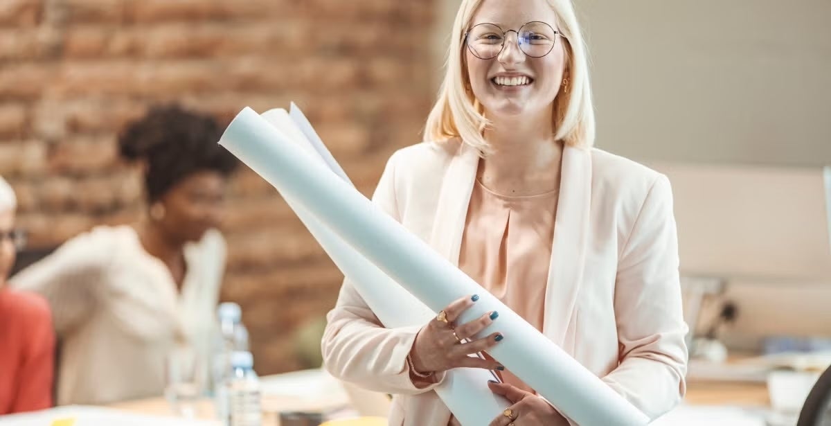 A cheerful woman holding blueprints, wearing glasses and a light blazer, stands in an office setting.